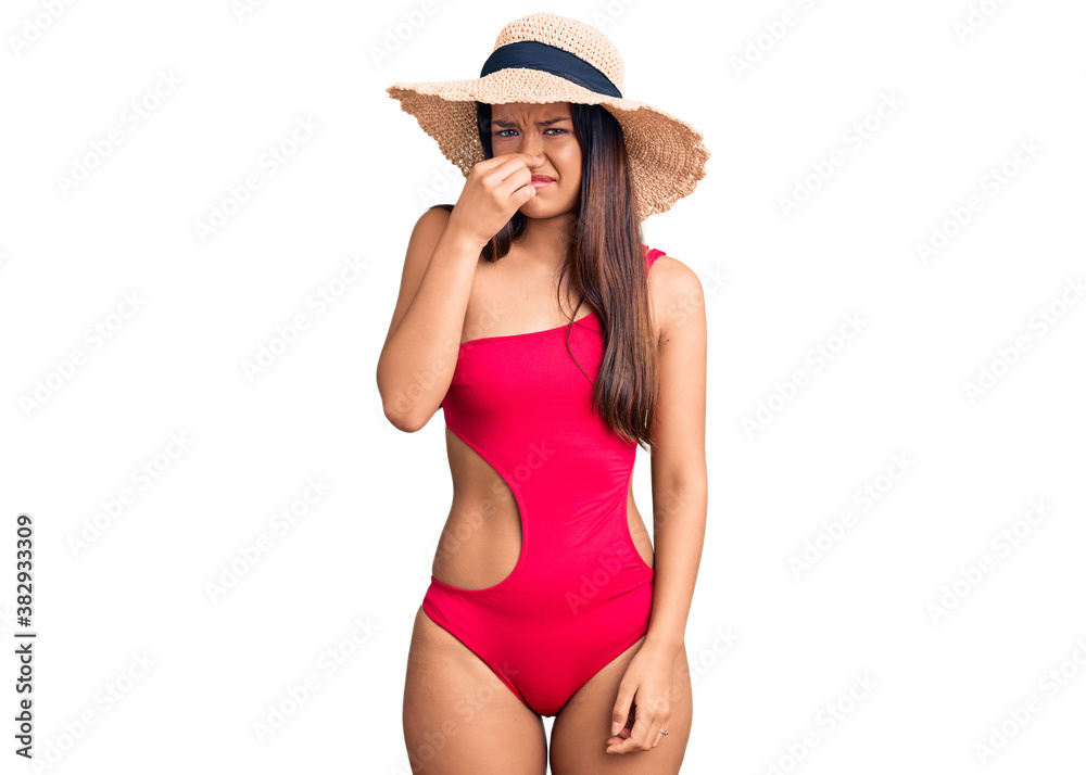 Young beautiful latin girl wearing swimwear and summer hat smelling something stinky and disgusting, intolerable smell, holding breath with fingers on nose. bad smell Young beautiful latin girl wearing swimwear and summer hat smelling something stinky and disgusting, intolerable smell, holding breath with fingers on nose. bad smell