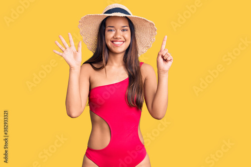 Young beautiful latin girl wearing swimwear and summer hat showing and pointing up with fingers number six while smiling confident and happy.