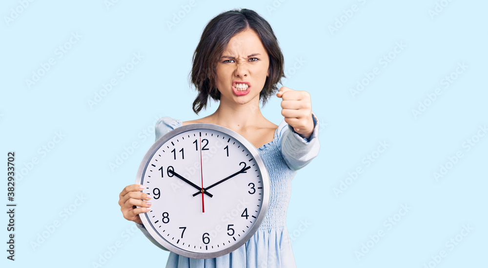 Young beautiful girl holding big clock annoyed and frustrated shouting with anger, yelling crazy with anger and hand raised Young beautiful girl holding big clock annoyed and frustrated shouting with anger, yelling crazy with anger and hand raised