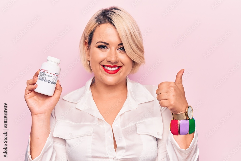 Young beautiful blonde plus size woman holding jar of pills over isolated pink background smiling happy and positive, thumb up doing excellent and approval sign Young beautiful blonde plus size woman holding jar of pills over isolated pink background smiling happy and positive, thumb up doing excellent and approval sign