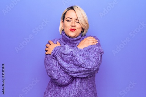 Beautiful blonde plus size woman wearing casual turtleneck sweater over purple background Hugging oneself happy and positive, smiling confident. Self love and self care © Krakenimages.com