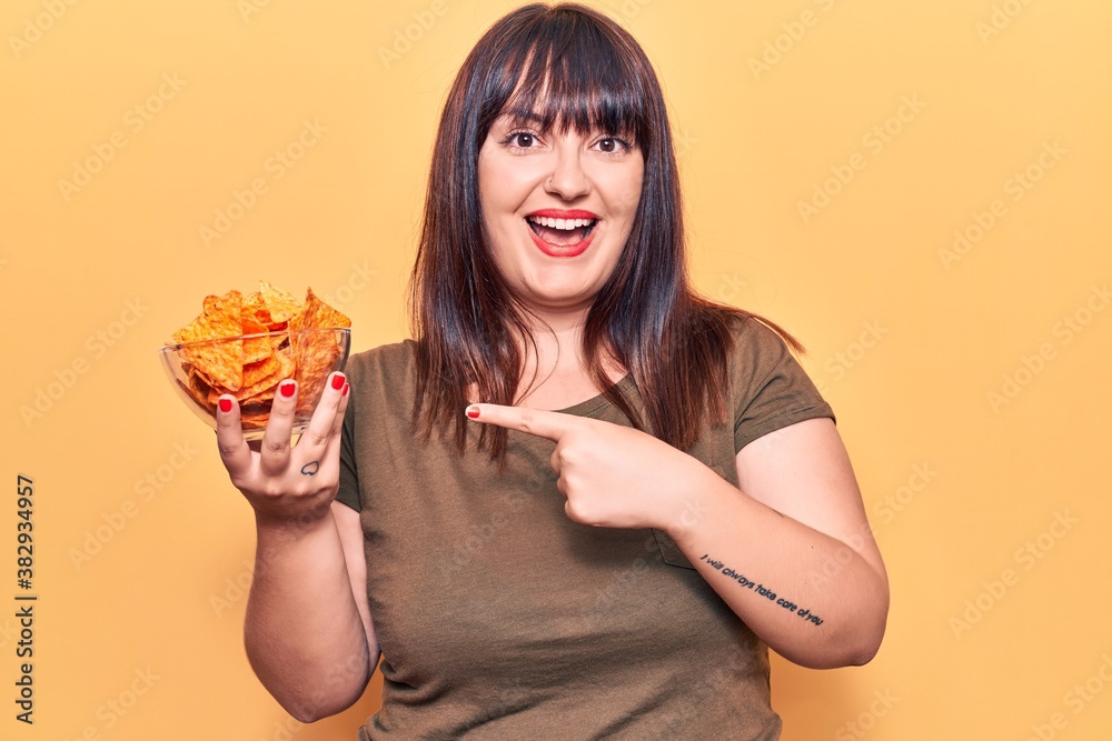 Young plus size woman holding nachos potato chips smiling happy pointing with hand and finger Young plus size woman holding nachos potato chips smiling happy pointing with hand and finger