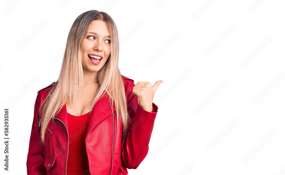 Young beautiful blonde woman wearing red leather jacket smiling with happy face looking and pointing to the side with thumb up. Young beautiful blonde woman wearing red leather jacket smiling with happy face looking and pointing to the side with thumb up.