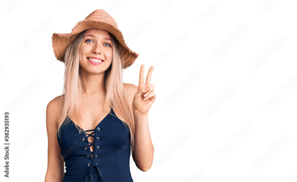 Young beautiful blonde woman wearing swimwear and summer hat smiling with happy face winking at the camera doing victory sign. number two.