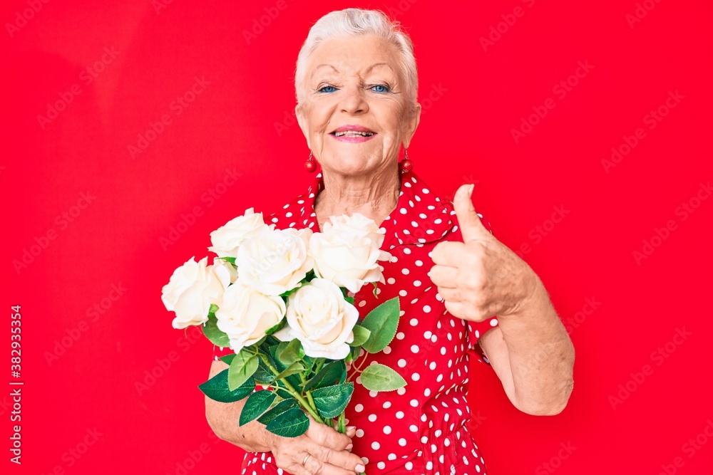 Senior beautiful woman with blue eyes and grey hair holding bouquet of white flowers smiling happy and positive, thumb up doing excellent and approval sign Senior beautiful woman with blue eyes and grey hair holding bouquet of white flowers smiling happy and positive, thumb up doing excellent and approval sign