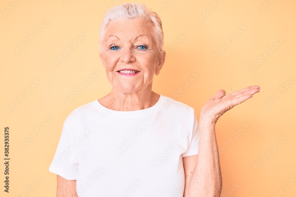 Senior beautiful woman with blue eyes and grey hair wearing classic white tshirt over yellow background smiling cheerful presenting and pointing with palm of hand looking at the camera. Senior beautiful woman with blue eyes and grey hair wearing classic white tshirt over yellow background smiling cheerful presenting and pointing with palm of hand looking at the camera.