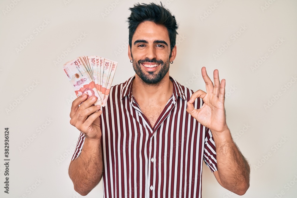 Handsome hispanic man with beard holding colombian pesos doing ok sign with fingers, smiling friendly gesturing excellent symbol Handsome hispanic man with beard holding colombian pesos doing ok sign with fingers, smiling friendly gesturing excellent symbol