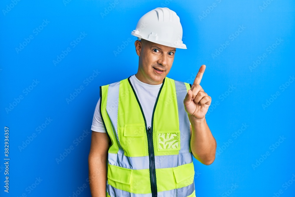 Middle age bald man wearing architect hardhat smiling with an idea or question pointing finger up with happy face, number one