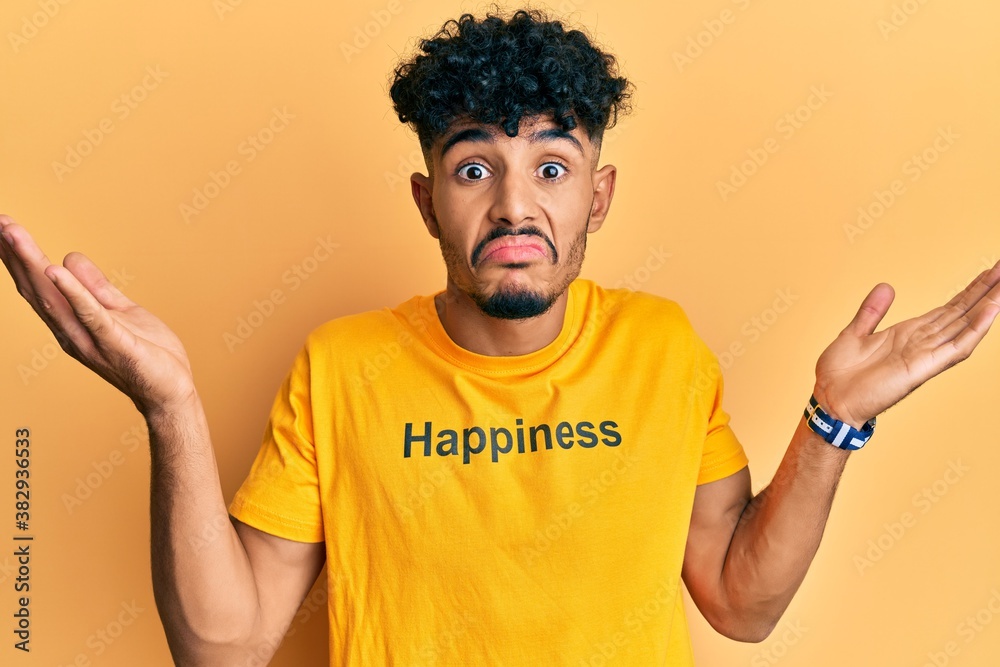 Young arab handsome man wearing tshirt with happiness word message clueless and confused with open arms, no idea and doubtful face. Young arab handsome man wearing tshirt with happiness word message clueless and confused with open arms, no idea and doubtful face.