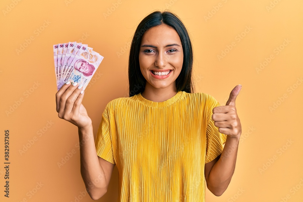 Young brunette woman holding 50 mexican pesos banknotes smiling happy and positive, thumb up doing excellent and approval sign Young brunette woman holding 50 mexican pesos banknotes smiling happy and positive, thumb up doing excellent and approval sign