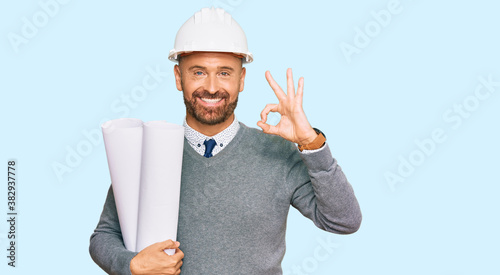 Handsome middle age man holding paper blueprints doing ok sign with fingers, smiling friendly gesturing excellent symbol photo