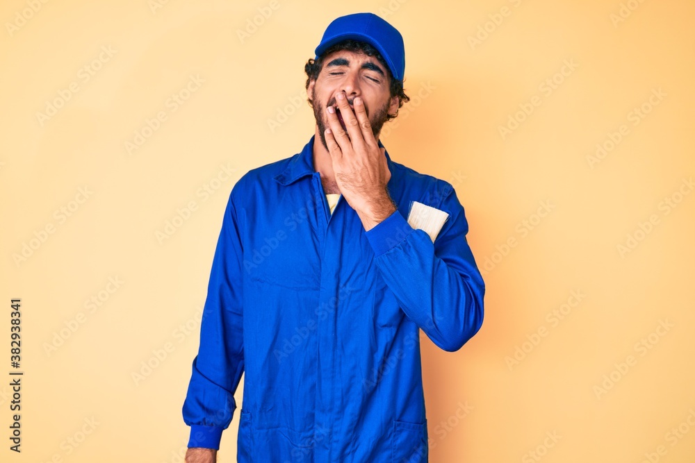 Handsome young man with curly hair and bear wearing builder jumpsuit uniform bored yawning tired covering mouth with hand. restless and sleepiness.