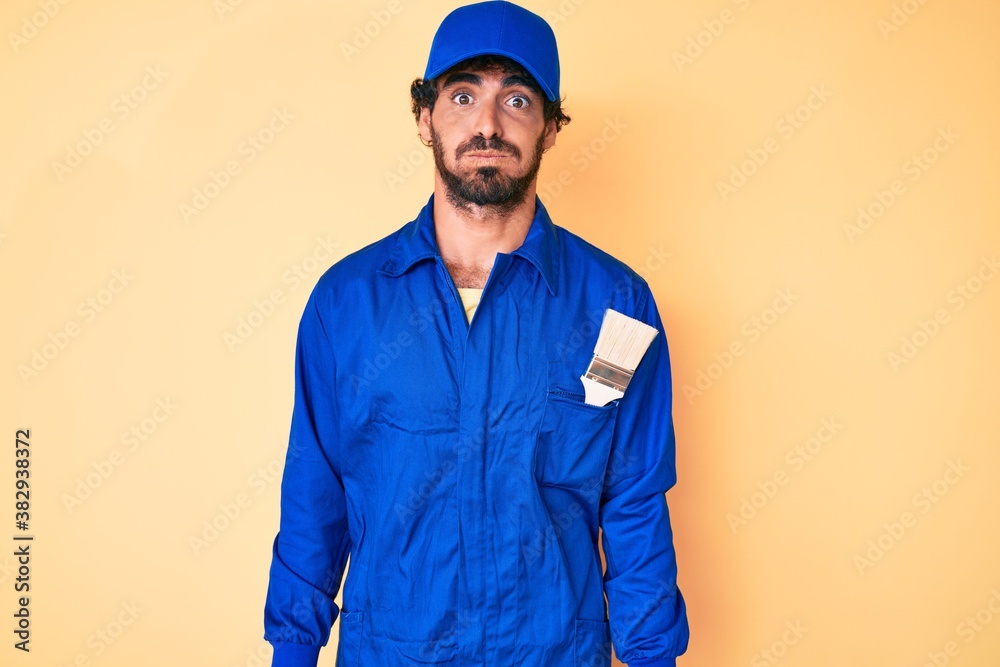 Handsome young man with curly hair and bear wearing builder jumpsuit uniform puffing cheeks with funny face. mouth inflated with air, crazy expression.