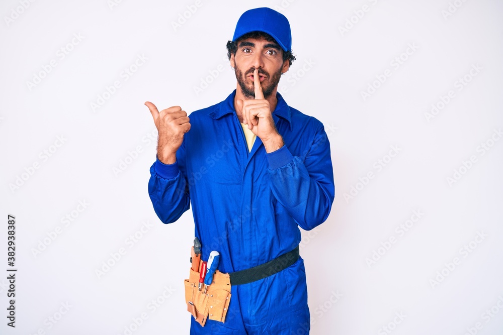 Handsome young man with curly hair and bear weaing handyman uniform asking to be quiet with finger on lips pointing with hand to the side. silence and secret concept.