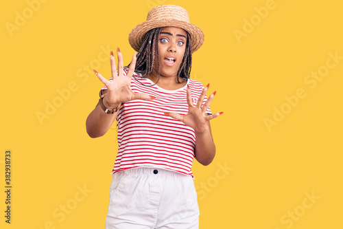 Young african american woman with braids wearing summer hat afraid and terrified with fear expression stop gesture with hands, shouting in shock. panic concept.