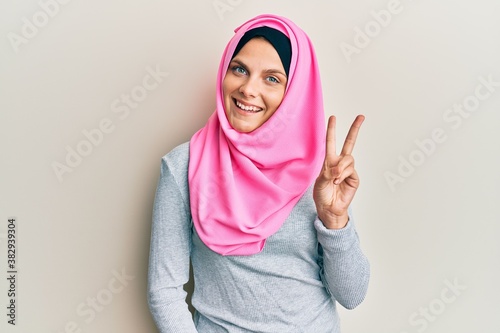 Young caucasian woman wearing traditional islamic hijab scarf smiling with happy face winking at the camera doing victory sign. number two.