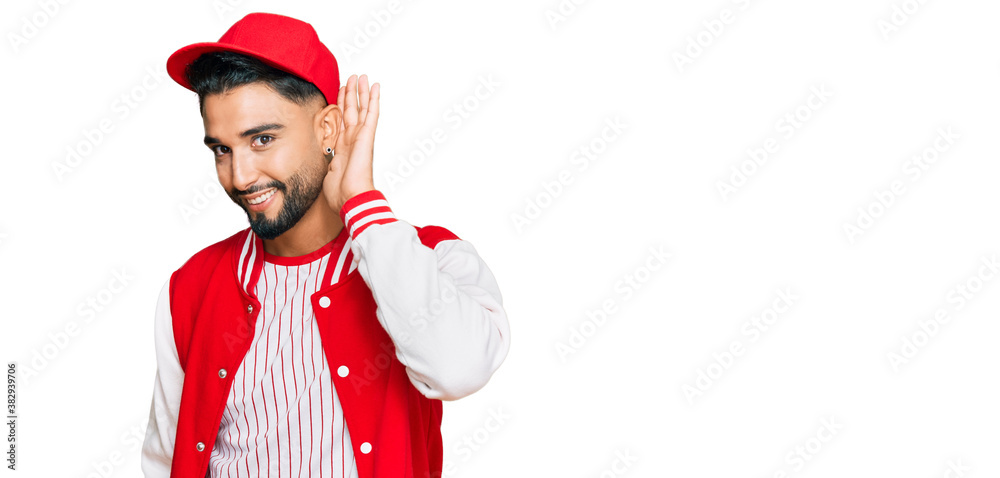 Young man with beard wearing baseball uniform smiling with hand over ear listening an hearing to rumor or gossip. deafness concept. Young man with beard wearing baseball uniform smiling with hand over ear listening an hearing to rumor or gossip. deafness concept.