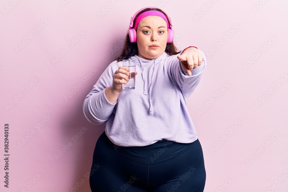 Young plus size woman drinking glass of water using headphones pointing with finger to the camera and to you, confident gesture looking serious Young plus size woman drinking glass of water using headphones pointing with finger to the camera and to you, confident gesture looking serious