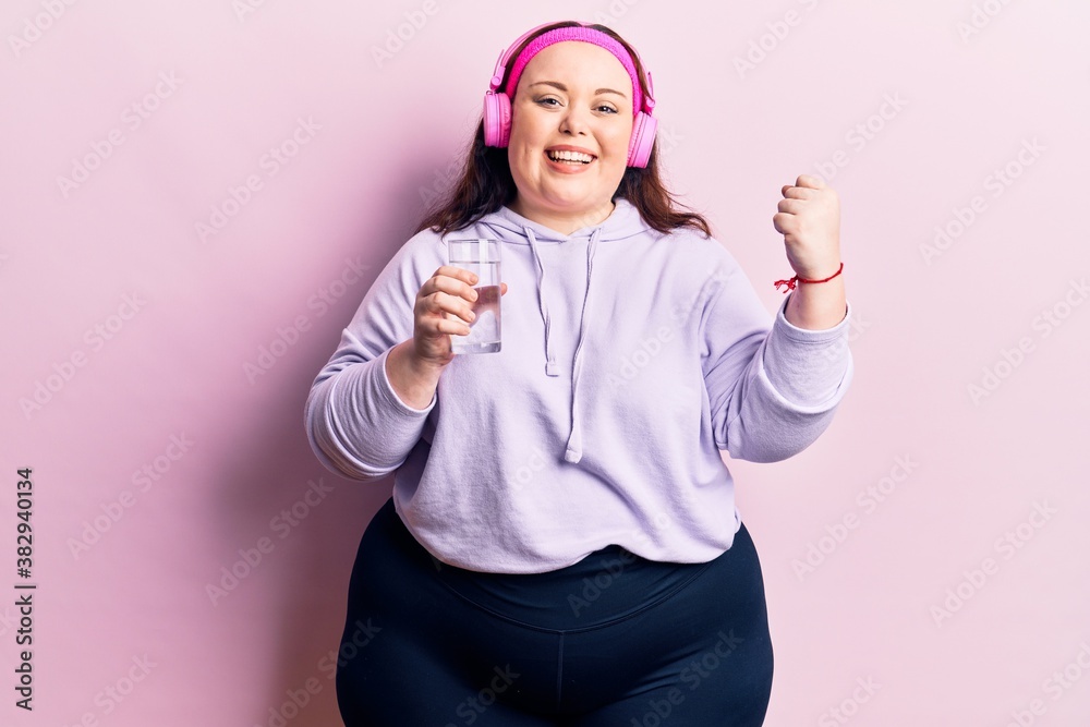 Naklejka premium Young plus size woman drinking glass of water using headphones screaming proud, celebrating victory and success very excited with raised arms