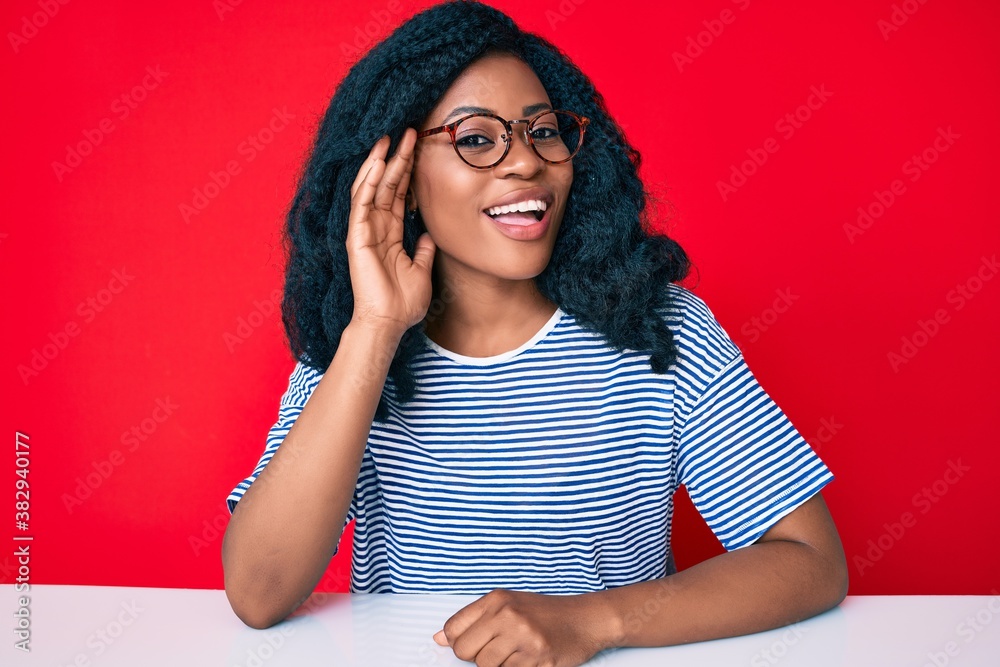 Beautiful african woman wearing casual clothes sitting on the table smiling with hand over ear listening and hearing to rumor or gossip. deafness concept. Beautiful african woman wearing casual clothes sitting on the table smiling with hand over ear listening and hearing to rumor or gossip. deafness concept.