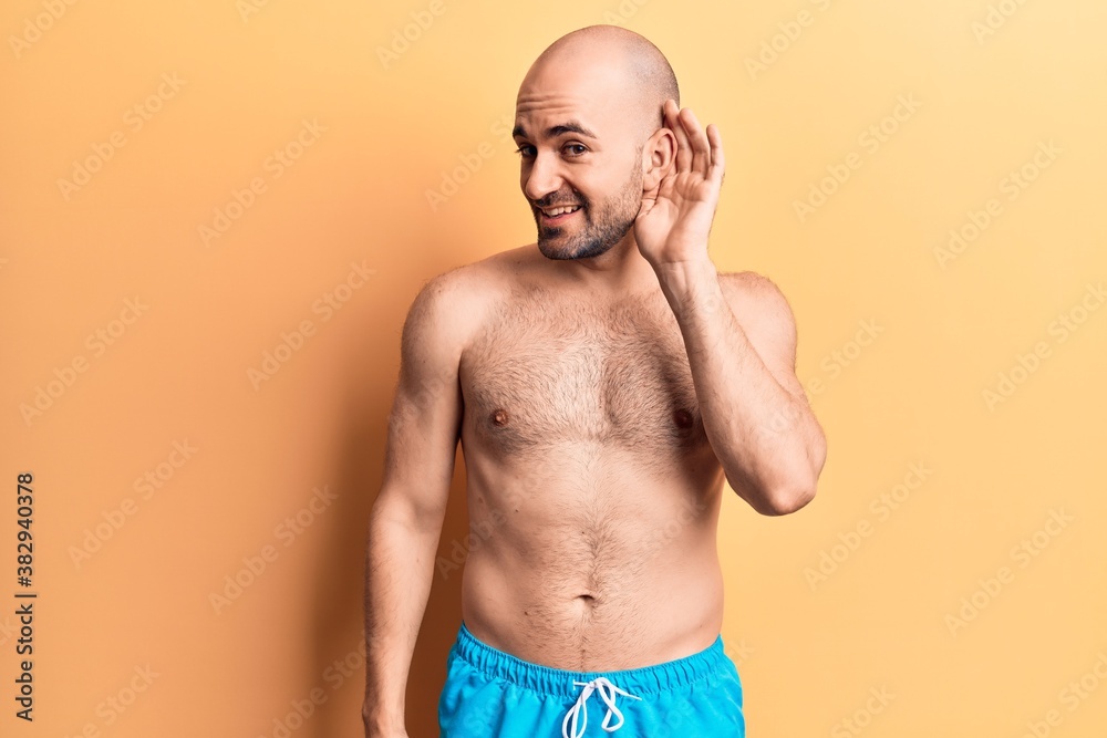 Young handsome bald man wearing swimwear shirtless smiling with hand over ear listening and hearing to rumor or gossip. deafness concept.