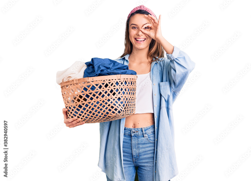 Young beautiful girl holding laundry basket smiling happy doing ok sign with hand on eye looking through fingers Young beautiful girl holding laundry basket smiling happy doing ok sign with hand on eye looking through fingers