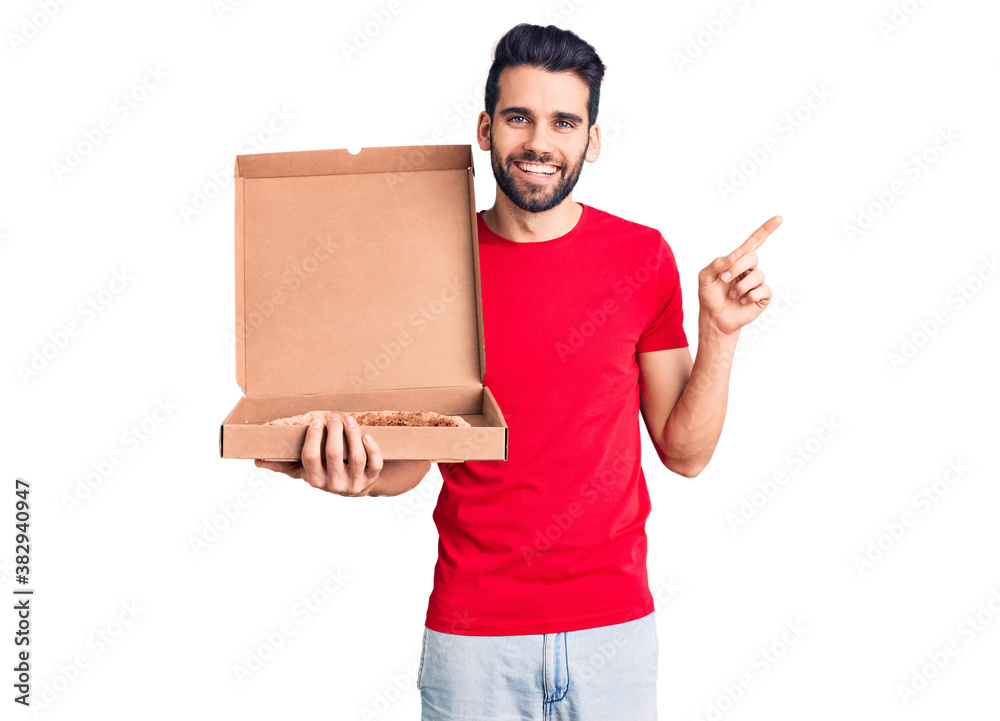 Young handsome man with beard holding delivery cardoboard with italian pizza smiling happy pointing with hand and finger to the side Young handsome man with beard holding delivery cardoboard with italian pizza smiling happy pointing with hand and finger to the side