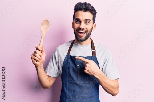 Young handsome man with beard wearing apron holding wooden spoon smiling happy pointing with hand and finger