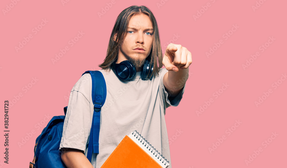 Handsome caucasian man with long hair wearing student backpack and holding books pointing with finger to the camera and to you, confident gesture looking serious