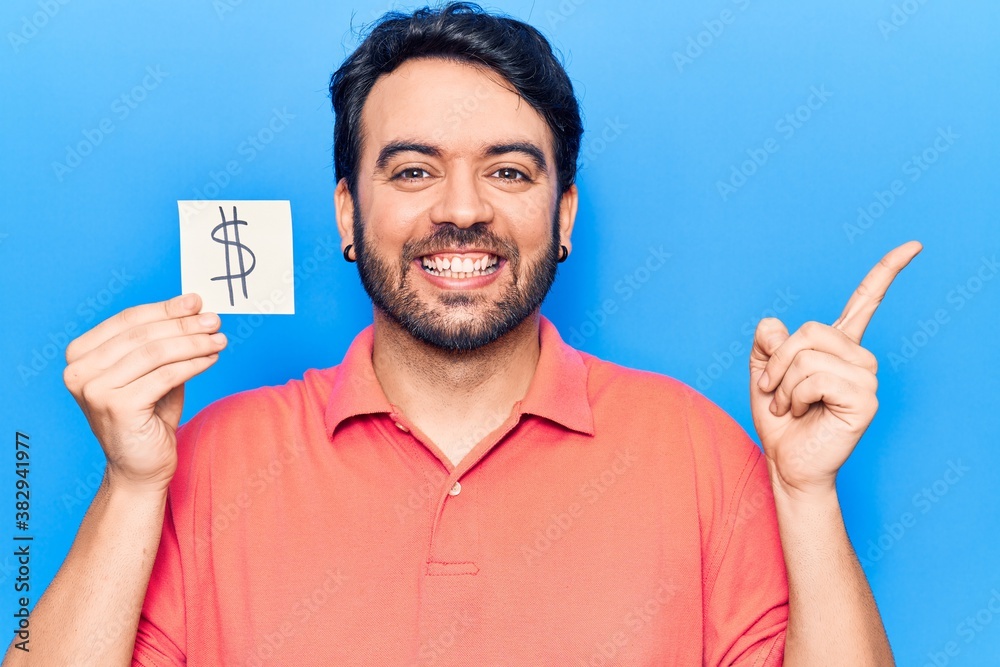Young hispanic man holding dollar symbol reminder paper smiling happy pointing with hand and finger to the side Young hispanic man holding dollar symbol reminder paper smiling happy pointing with hand and finger to the side