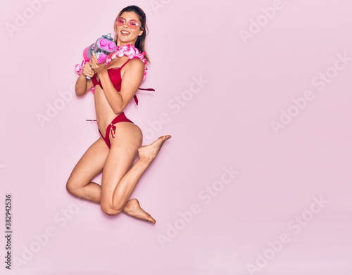 Young beautiful girl on vacation wearing bikini and hawaiian lei smiling happy. Jumping with smile on face holding water gun over isolated pink background