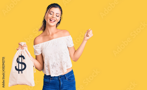Young beautiful woman holding money bag with dollar symbol screaming proud  celebrating victory and success very excited with raised arms