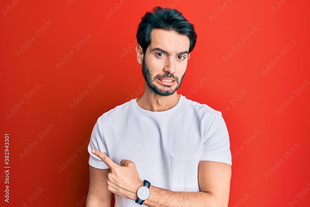 Young hispanic man wearing casual white tshirt pointing aside worried and nervous with forefinger, concerned and surprised expression Young hispanic man wearing casual white tshirt pointing aside worried and nervous with forefinger, concerned and surprised expression
