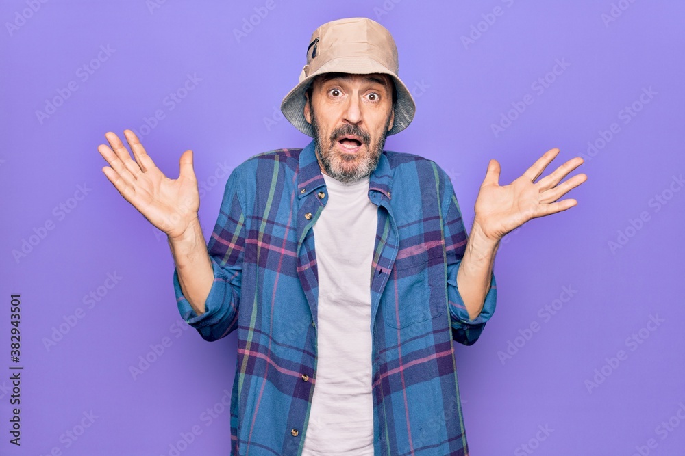 Middle age handsome man wearing casual shirt and hat over isolated purple background clueless and confused with open arms, no idea and doubtful face.
