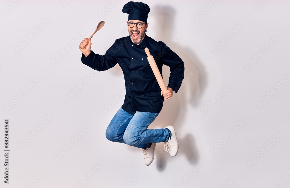 Middle age handsome hispanic man wearing glasses and cook uniform smiling happy. Jumping with smile on face holding kneader and spoon over isolated white background