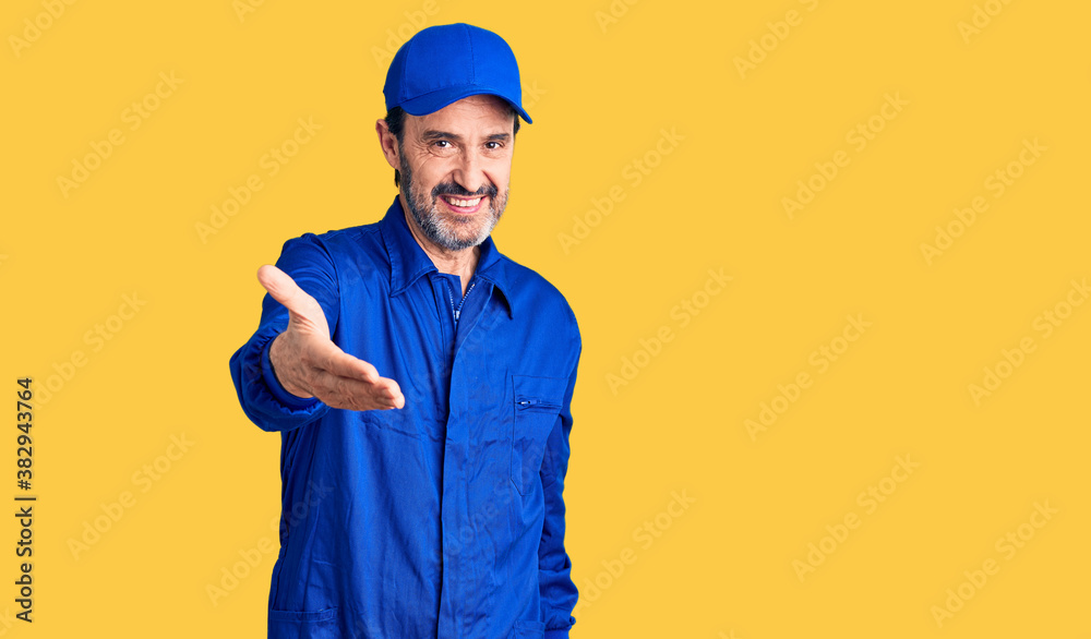 Middle age handsome man wearing mechanic uniform smiling cheerful offering palm hand giving assistance and acceptance.