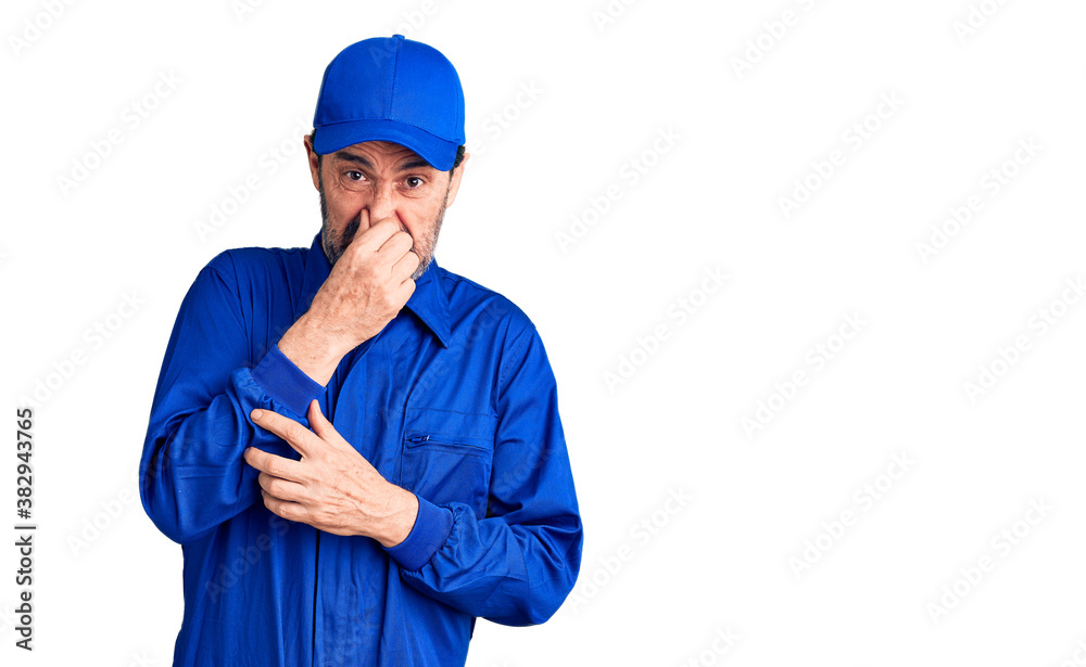 Middle age handsome man wearing mechanic uniform smelling something stinky and disgusting, intolerable smell, holding breath with fingers on nose. bad smell Middle age handsome man wearing mechanic uniform smelling something stinky and disgusting, intolerable smell, holding breath with fingers on nose. bad smell