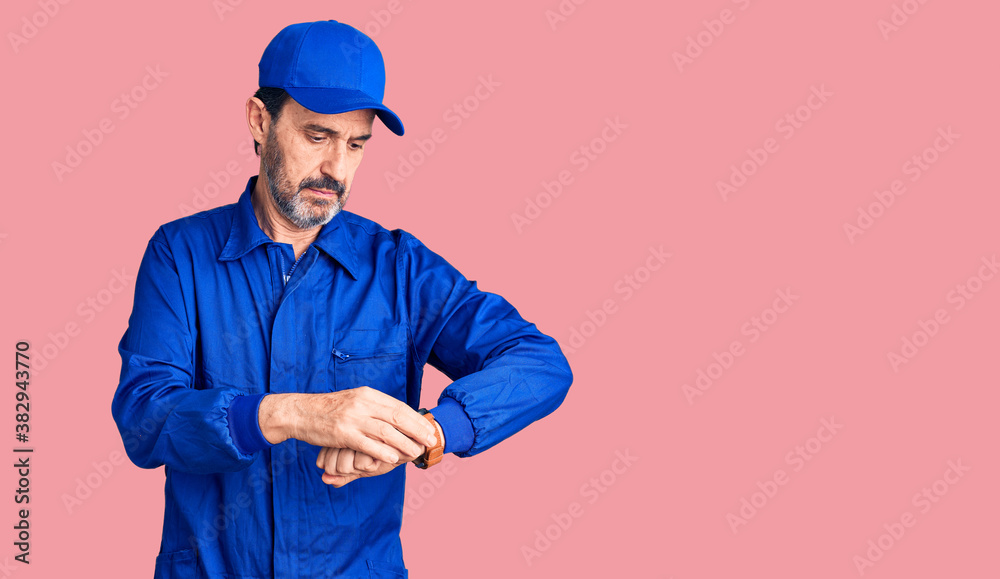 Middle age handsome man wearing mechanic uniform checking the time on wrist watch, relaxed and confident