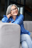 Happy relaxed mature old adult woman wearing glasses resting sitting on couch at home. Smiling mid age grey-haired elegant senior lady relaxing on comfortable sofa looking at camera. Vertical portrait