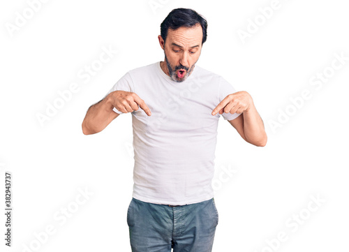 Middle age handsome man wearing casual t-shirt pointing down with fingers showing advertisement, surprised face and open mouth