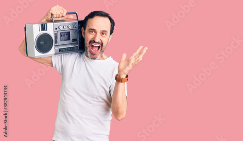 Middle age handsome man listening to music using vintage boombox celebrating victory with happy smile and winner expression with raised hands