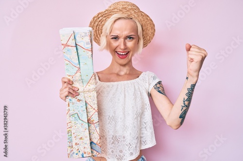 Young blonde woman wearing summer hat holding city map screaming proud, celebrating victory and success very excited with raised arms © Krakenimages.com