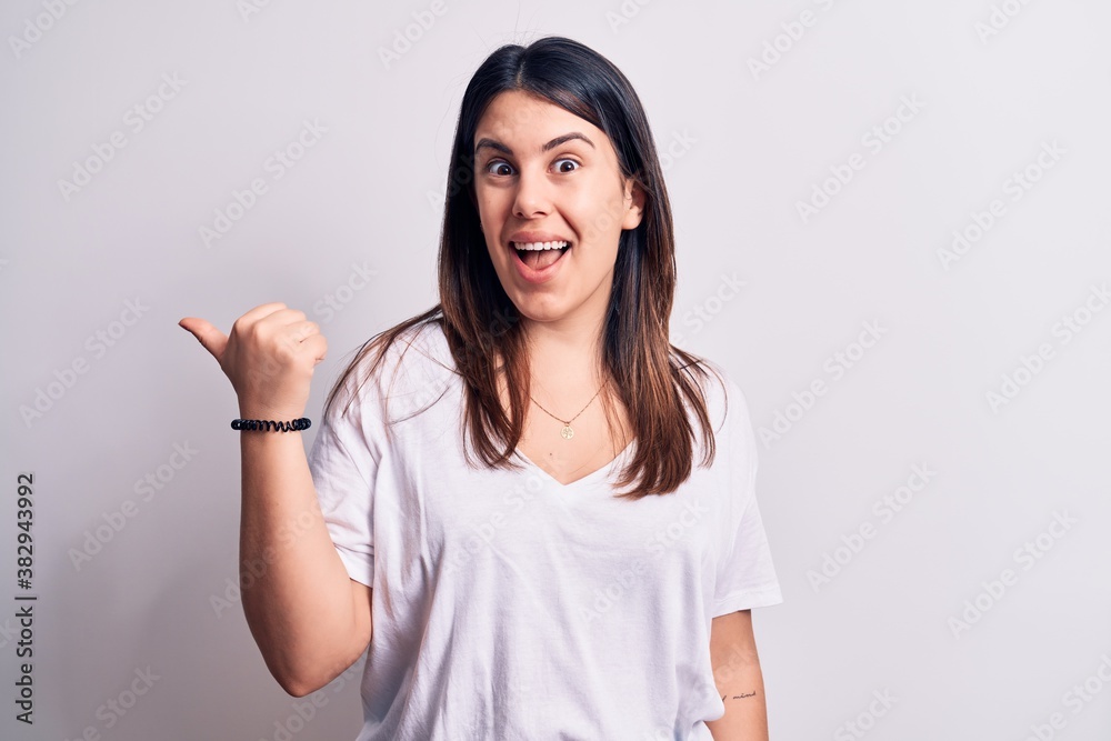 Young beautiful brunette woman wearing casual t-shirt standing over isolated white background pointing thumb up to the side smiling happy with open mouth Young beautiful brunette woman wearing casual t-shirt standing over isolated white background pointing thumb up to the side smiling happy with open mouth