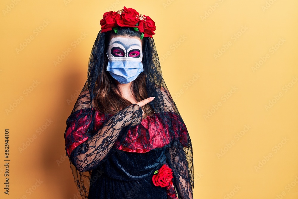 Young woman wearing day of the dead costume wearing medical mask smiling happy pointing with hand and finger to the side