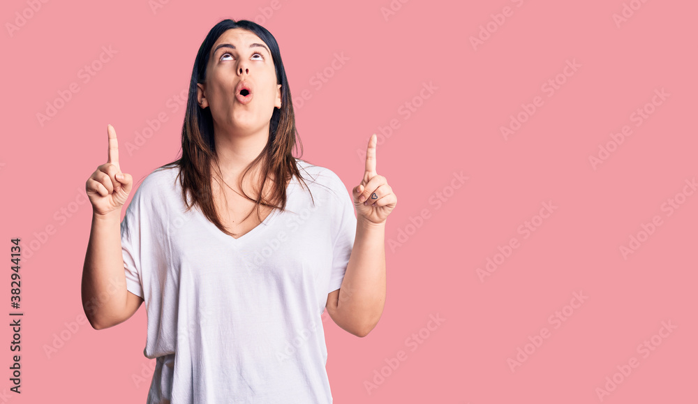 Young beautiful brunette woman wearing casual t-shirt amazed and surprised looking up and pointing with fingers and raised arms. Young beautiful brunette woman wearing casual t-shirt amazed and surprised looking up and pointing with fingers and raised arms.