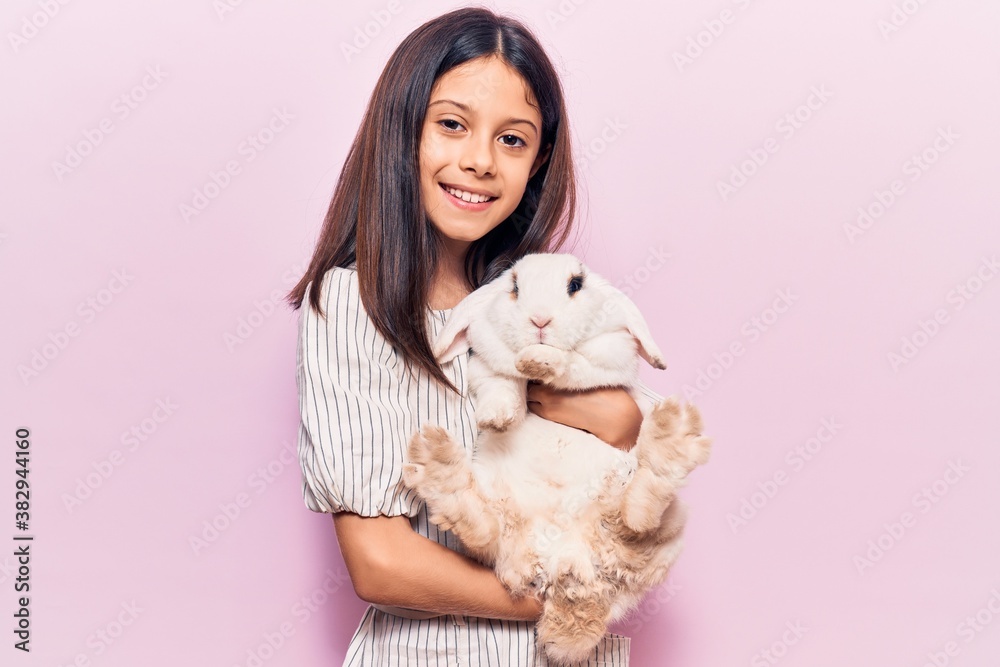 Adorable hispanic kid girl smiling happy. Holding cute rabbit standing over isolated pink background Adorable hispanic kid girl smiling happy. Holding cute rabbit standing over isolated pink background