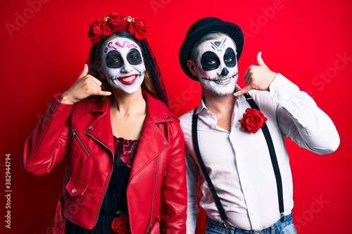 Couple wearing day of the dead costume over red smiling doing phone gesture with hand and fingers like talking on the telephone. communicating concepts.