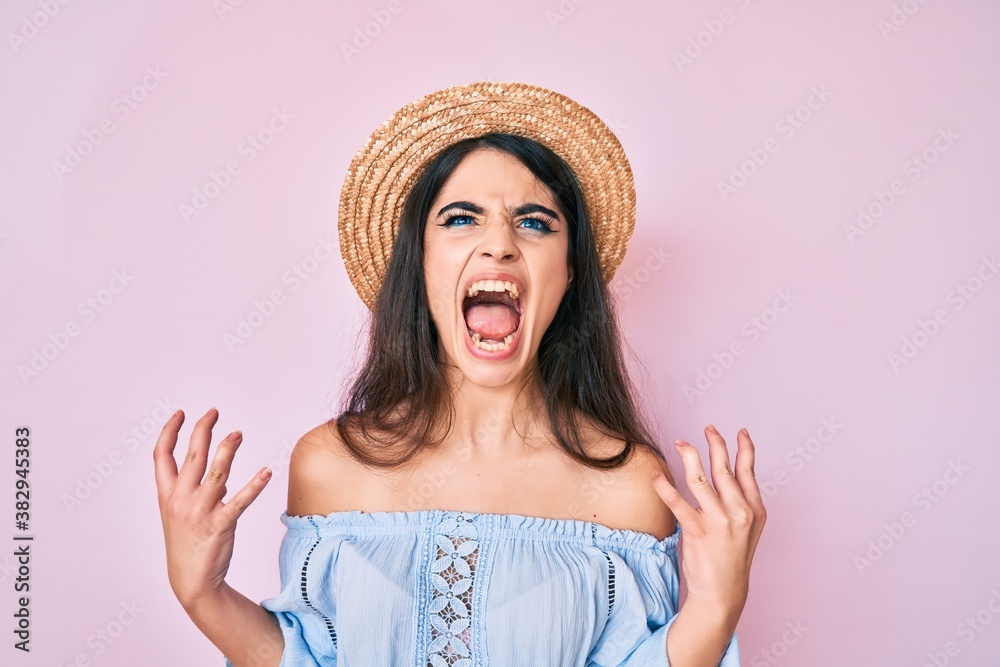 Brunette teenager girl wearing summer hat crazy and mad shouting and yelling with aggressive expression and arms raised. frustration concept. Brunette teenager girl wearing summer hat crazy and mad shouting and yelling with aggressive expression and arms raised. frustration concept.