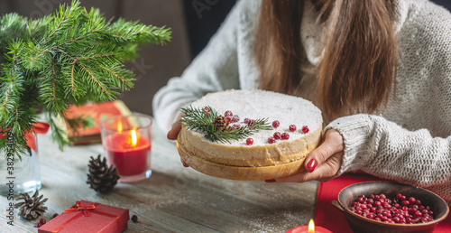 Woman is holding a festive white mousse cake covered with coconut flakes imitating snow and decorated with red berries, cones and fir branches. Concept of the New year and Christmas