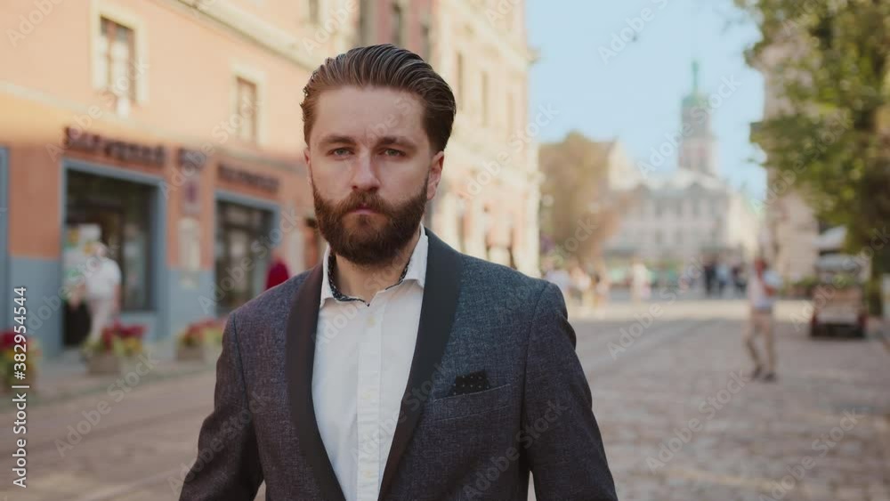 Portrait handsome bearded businessman standing look at camera serious looking at his watch in old city at sunlight. Elegant confident man. Entrepreneur leader successful. Slow motion Portrait handsome bearded businessman standing look at camera serious looking at his watch in old city at sunlight. Elegant confident man. Entrepreneur leader successful. Slow motion